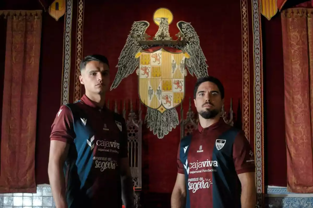 Dos futbolistas posan con uniforme granate y azul oscuro frente a un gran escudo heráldico en una sala decorada con cortinas rojas y detalles dorados