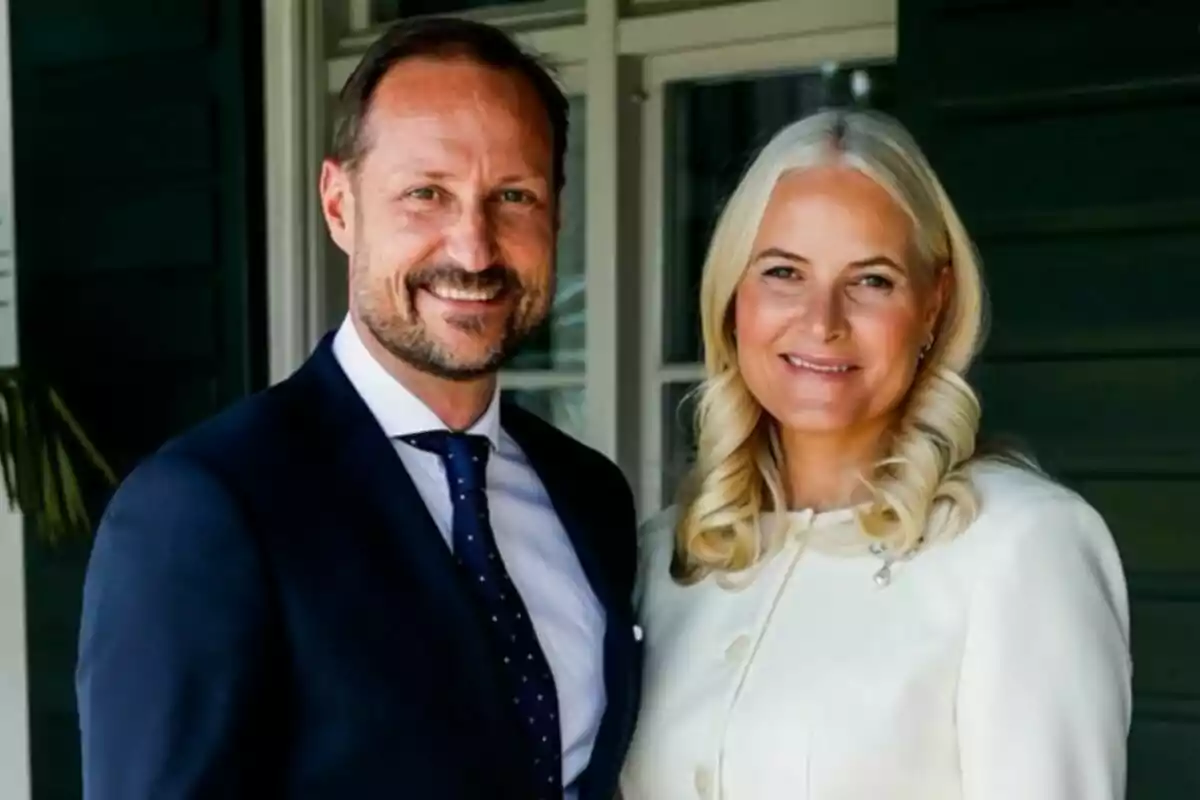 Una pareja sonriente posando juntos, el hombre lleva traje azul y la mujer viste de blanco, ambos están de pie frente a una casa Una pareja sonriente posando juntos, el hombre lleva traje azul y la mujer viste de blanco, ambos están de pie frente a una casa
