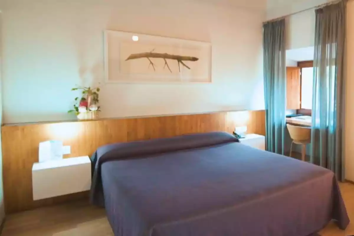 Habitación de hotel con cama grande, cabecera de madera, dos mesas flotantes blancas, lámparas encendidas, cuadro decorativo en la pared, cortinas azules y escritorio junto a la ventana Habitación de hotel con cama grande, cabecera de madera, dos mesas flotantes blancas, lámparas encendidas, cuadro decorativo en la pared, cortinas azules y escritorio junto a la ventana