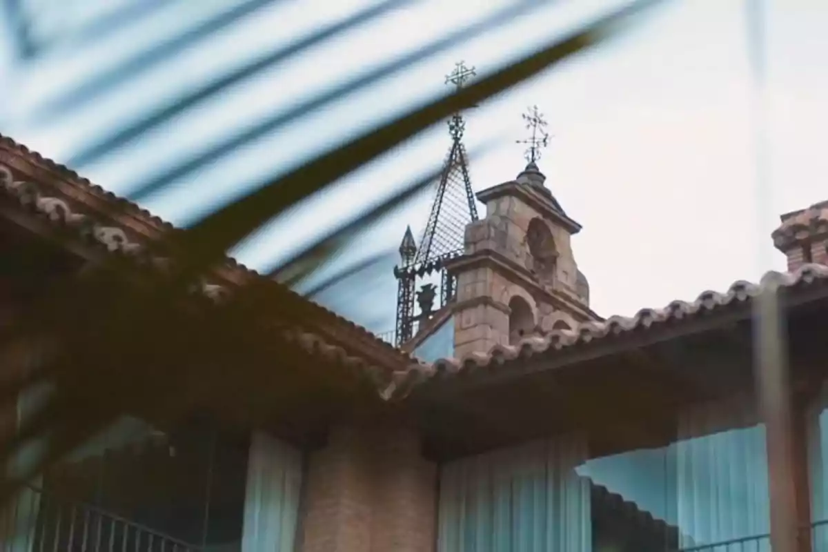 Campanario de piedra con detalles de hierro y cruces en la parte superior visto detrás de ramas y techos de tejas Campanario de piedra con detalles de hierro y cruces en la parte superior visto detrás de ramas y techos de tejas
