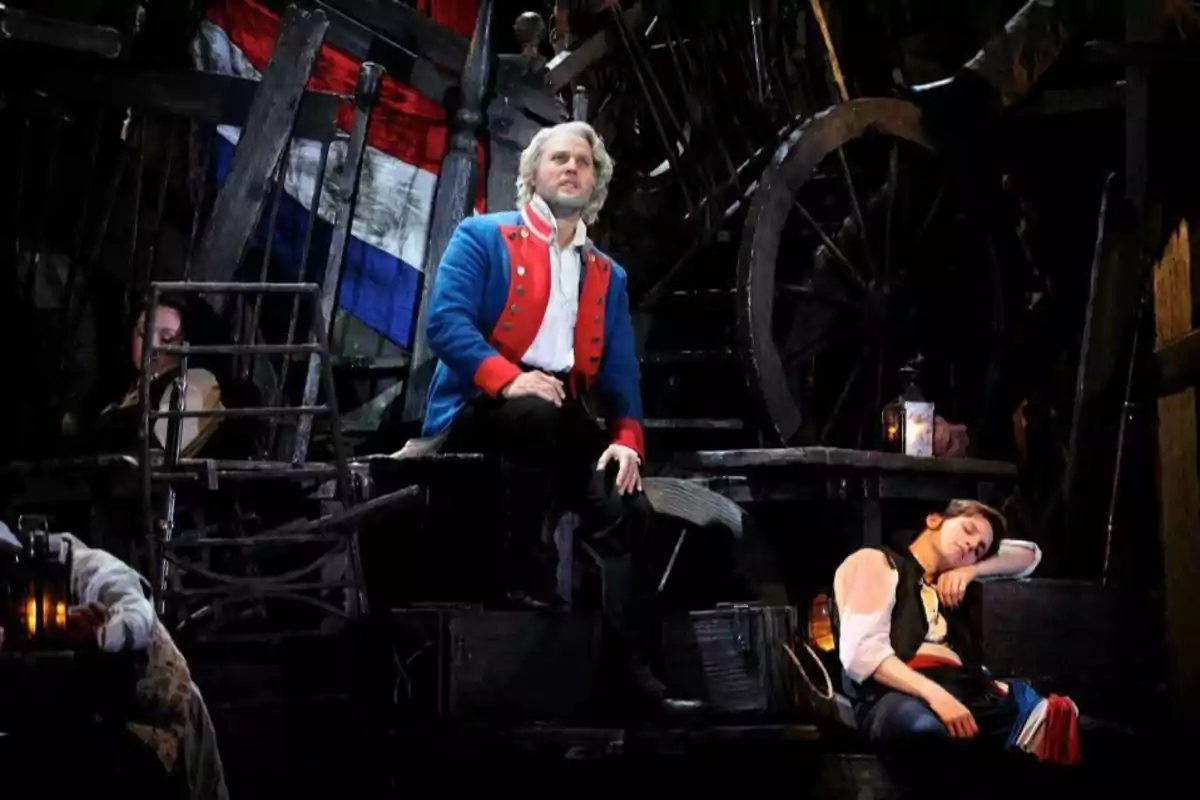 Escena teatral con un hombre de cabello canoso y chaqueta azul sentado en una barricada mientras otra persona descansa a su lado y al fondo se ve una bandera tricolor