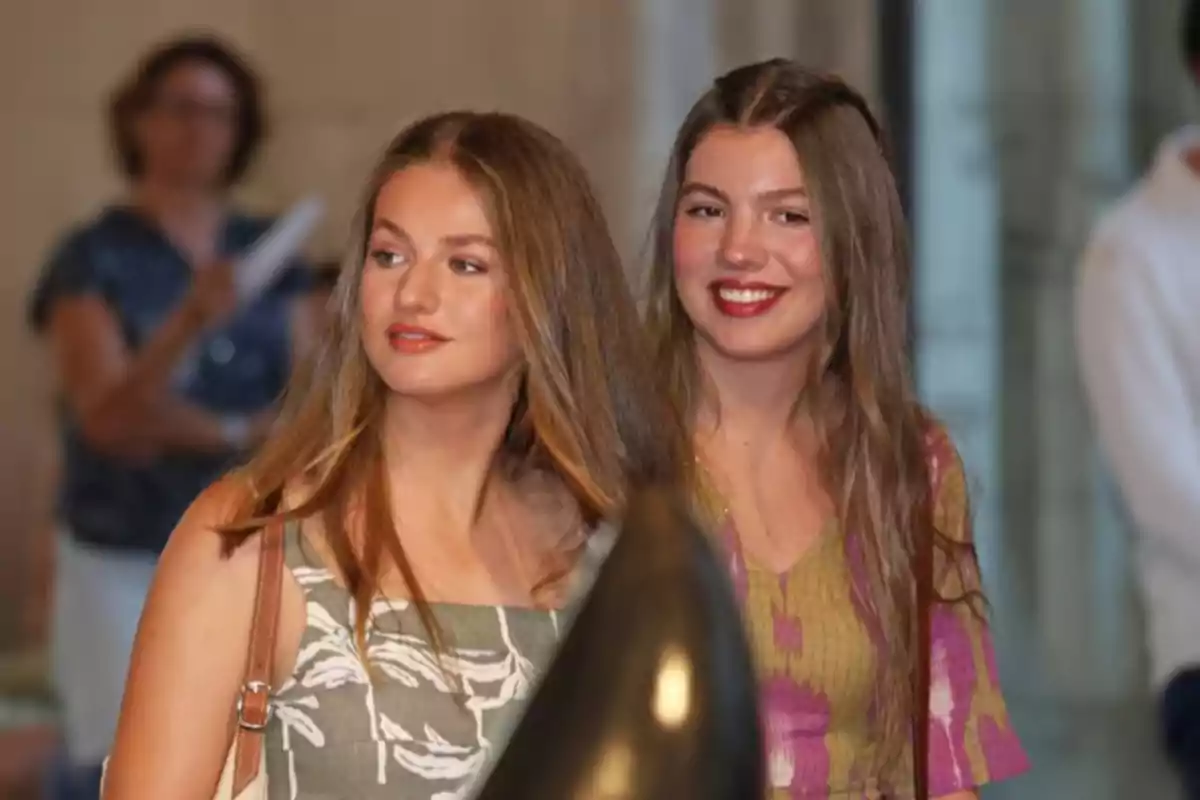 Two young girls with long, loose hair are standing side by side indoors, both wearing summer dresses and smiling while looking in different directions. Two young girls with long, loose hair are standing side by side indoors, both wearing summer dresses and smiling while looking in different directions.