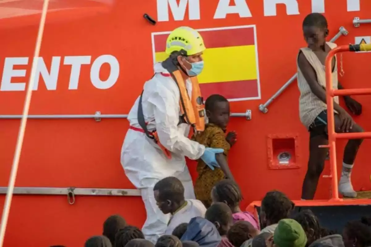 Rescatista con equipo de protección ayuda a un grupo de niños migrantes a bordo de una embarcación de Salvamento Marítimo de España