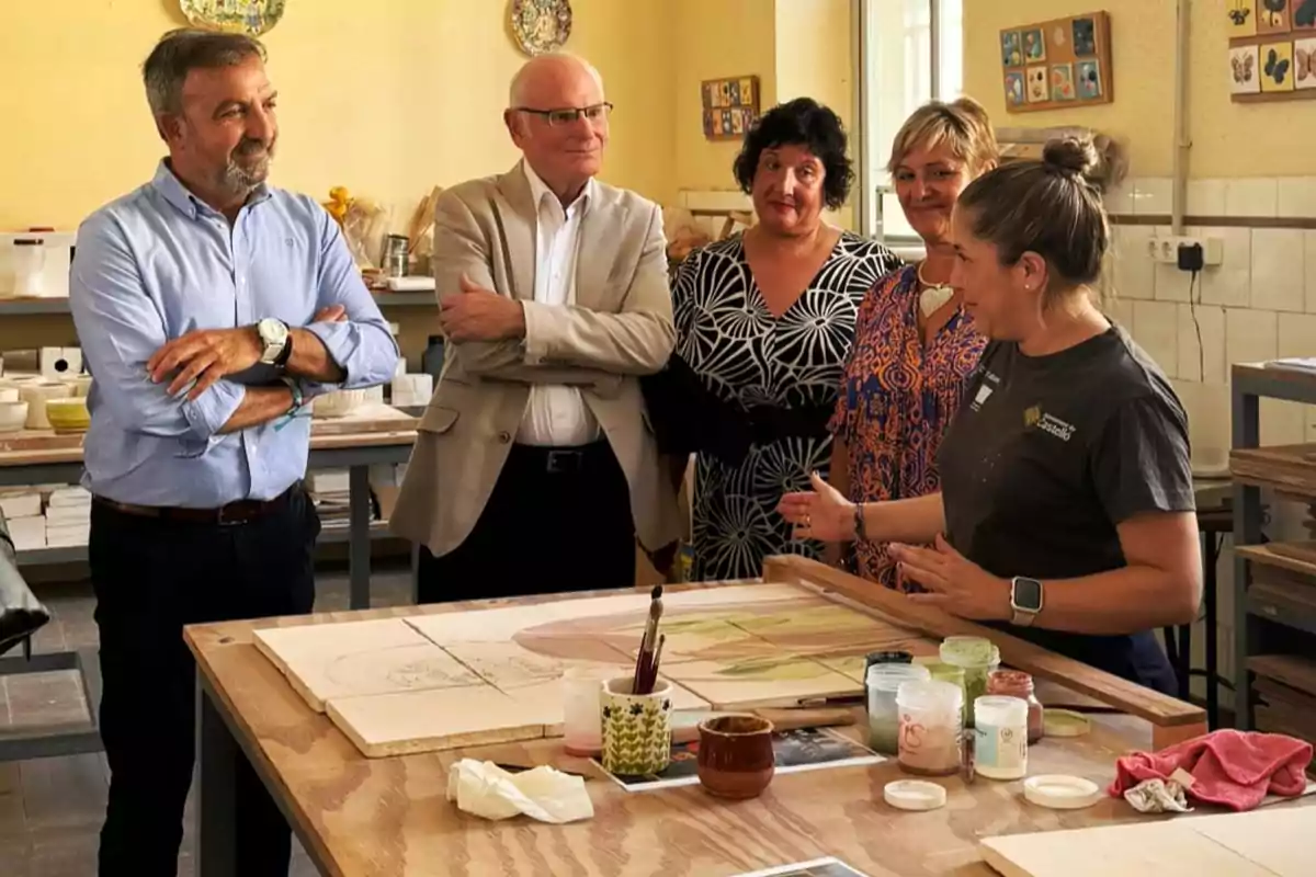 Cinco personas conversan en un taller de arte mientras una de ellas explica una técnica de pintura sobre azulejos en una mesa llena de materiales.