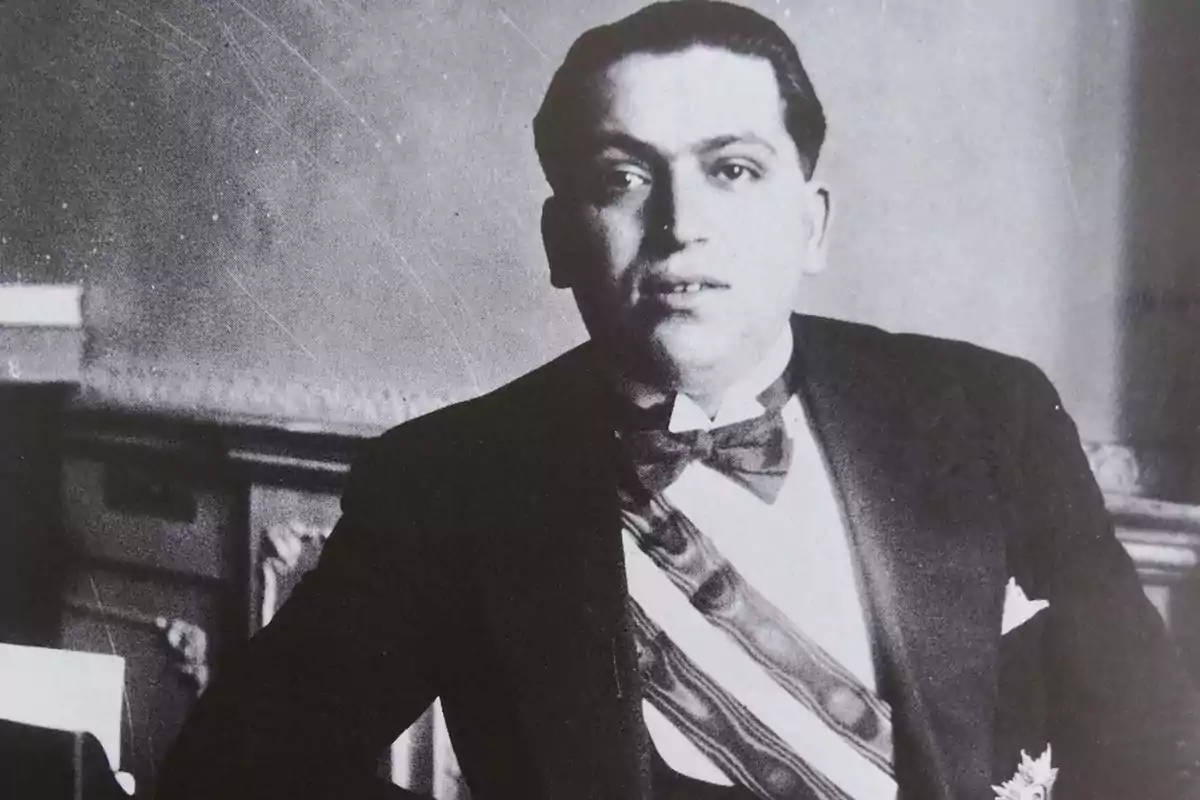 Hombre joven con esmoquin y banda presidencial sentado en una oficina antigua