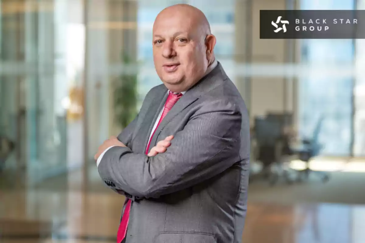Hombre calvo con traje gris y corbata roja posando con los brazos cruzados en una oficina moderna con el logo de Black Star Group en la esquina superior derecha