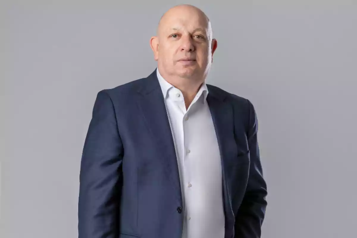 Hombre calvo de mediana edad vistiendo saco azul y camisa blanca posando frente a un fondo gris