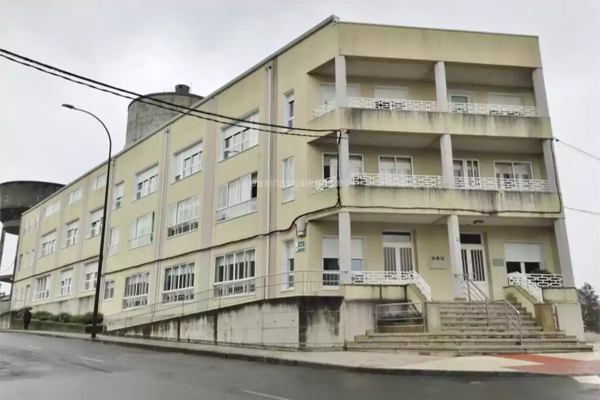 Edificio de tres plantas de color beige con varias ventanas y balcones, situado en una calle en pendiente y con un día nublado