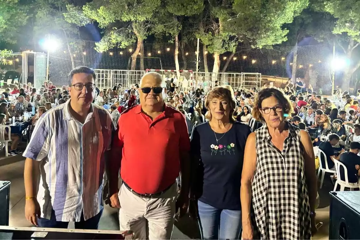 Cuatro personas posan al frente de un evento al aire libre por la noche con muchas personas sentadas en mesas y árboles iluminados al fondo