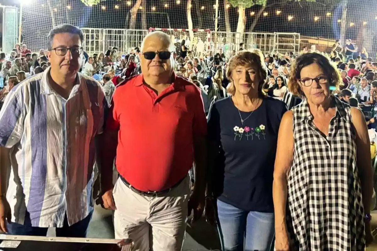 Cuatro personas posan al frente de un evento al aire libre con muchas personas sentadas al fondo y luces decorativas en la noche Cuatro personas posan al frente de un evento al aire libre con muchas personas sentadas al fondo y luces decorativas en la noche