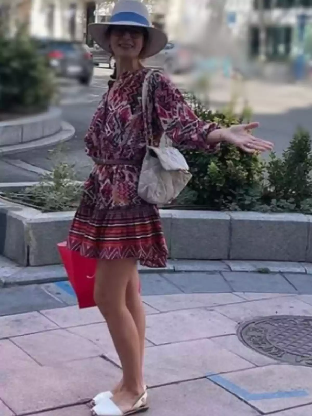 Smiling woman with a hat and patterned dress posing on an urban street with a white purse and a red shopping bag Smiling woman with a hat and patterned dress posing on an urban street with a white purse and a red shopping bag