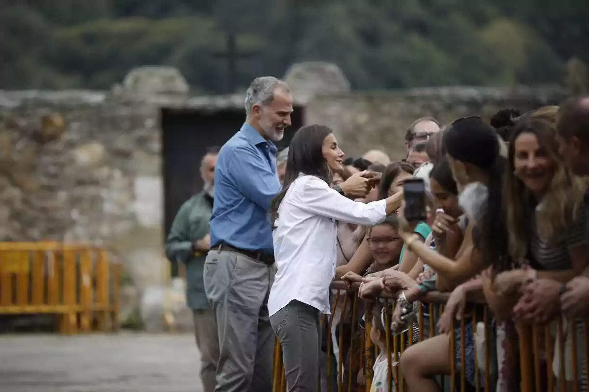 Una pareja saluda y conversa con un grupo de personas tras una valla en un entorno al aire libre con fondo de piedra y vegetación