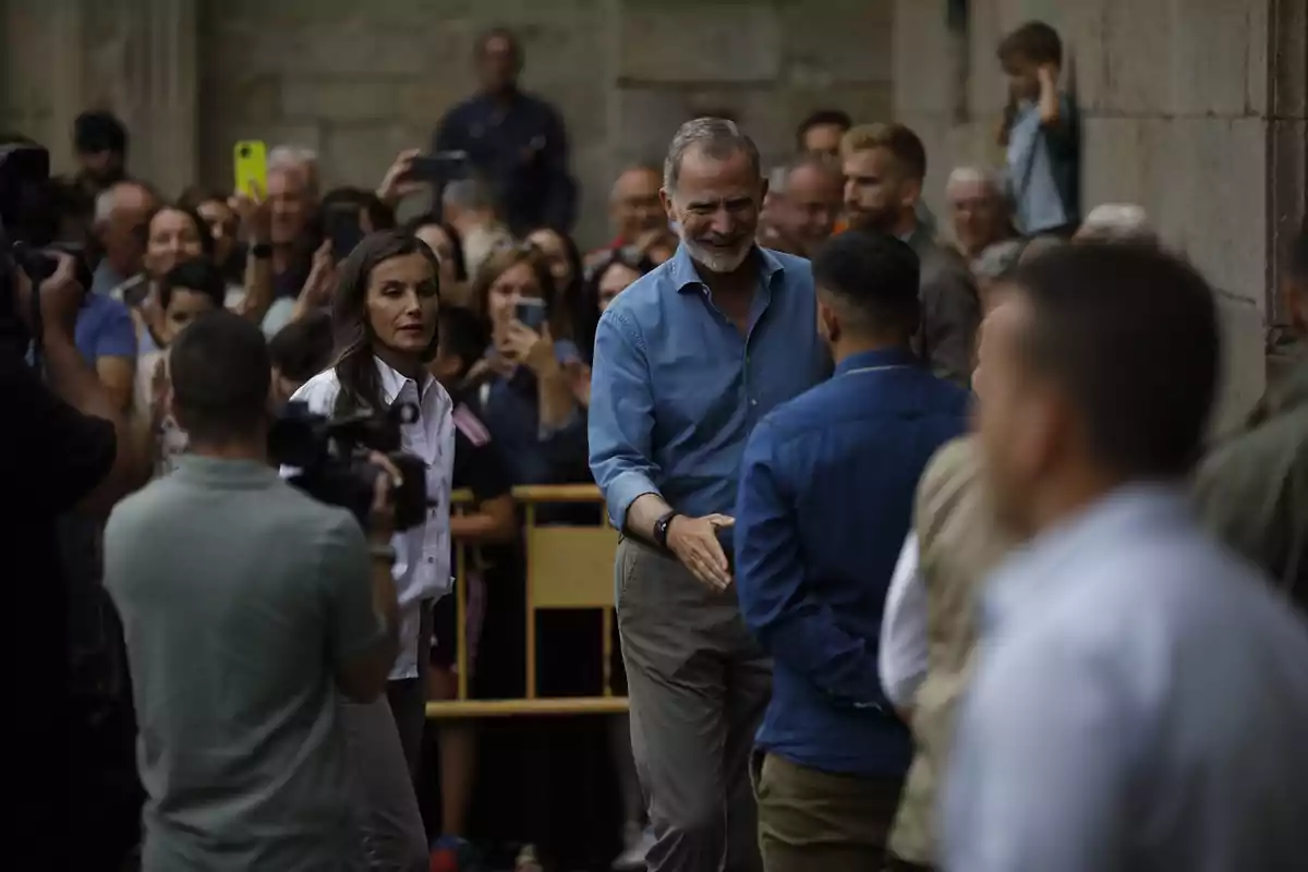 Un hombre sonriente saluda a otra persona mientras una mujer y varias personas observan y toman fotos en un evento al aire libre