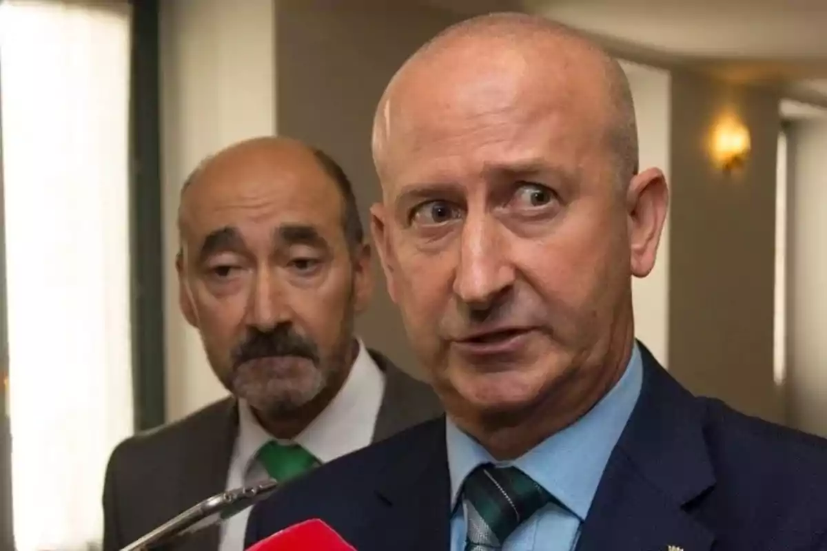 Dos hombres de traje en una entrevista, uno al frente hablando y el otro al fondo observando Dos hombres de traje en una entrevista, uno al frente hablando y el otro al fondo observando