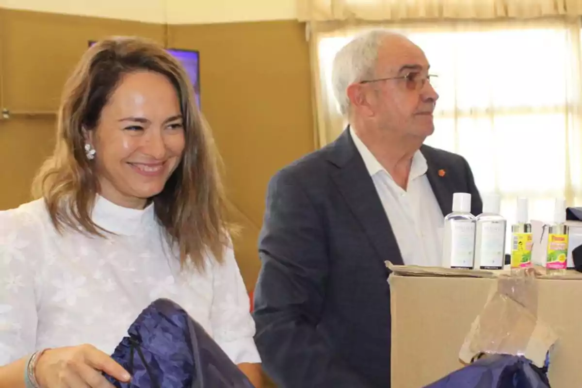 Una mujer sonriente y un hombre de cabello canoso están junto a una caja de cartón con botellas de gel antibacterial y otros productos en un ambiente interior iluminado. Una mujer sonriente y un hombre de cabello canoso están junto a una caja de cartón con botellas de gel antibacterial y otros productos en un ambiente interior iluminado.