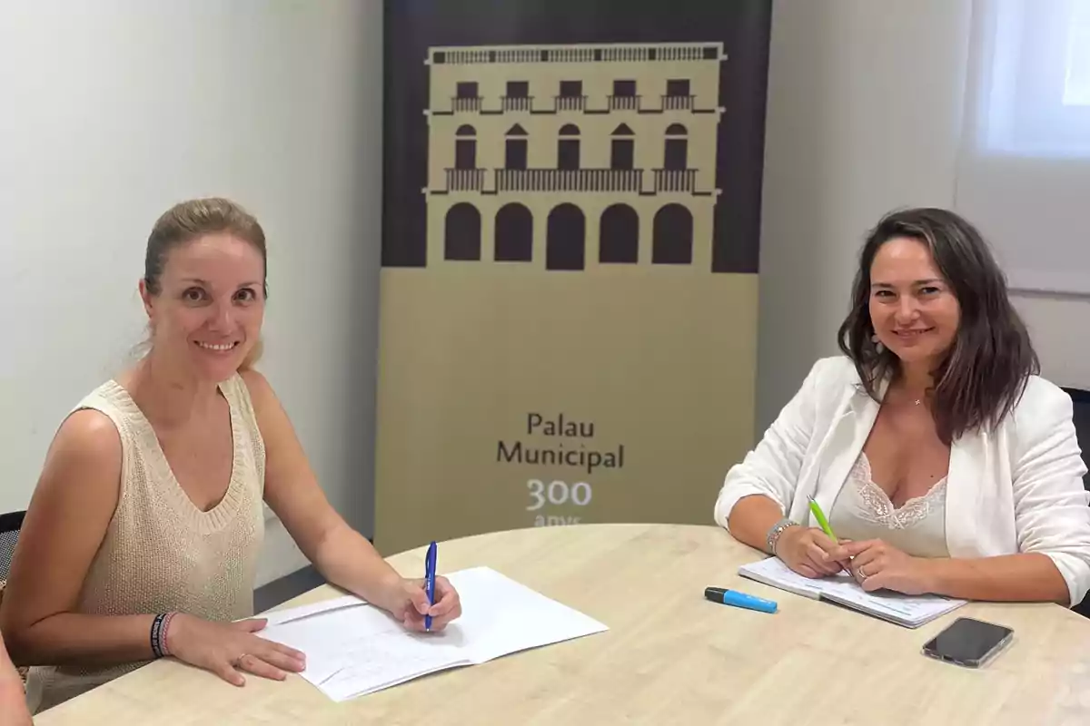 Dos mujeres sonríen mientras están sentadas en una mesa firmando documentos, al fondo hay un cartel con la imagen de un edificio y el texto Palau Municipal 300 anys. Dos mujeres sonríen mientras están sentadas en una mesa firmando documentos, al fondo hay un cartel con la imagen de un edificio y el texto Palau Municipal 300 anys.