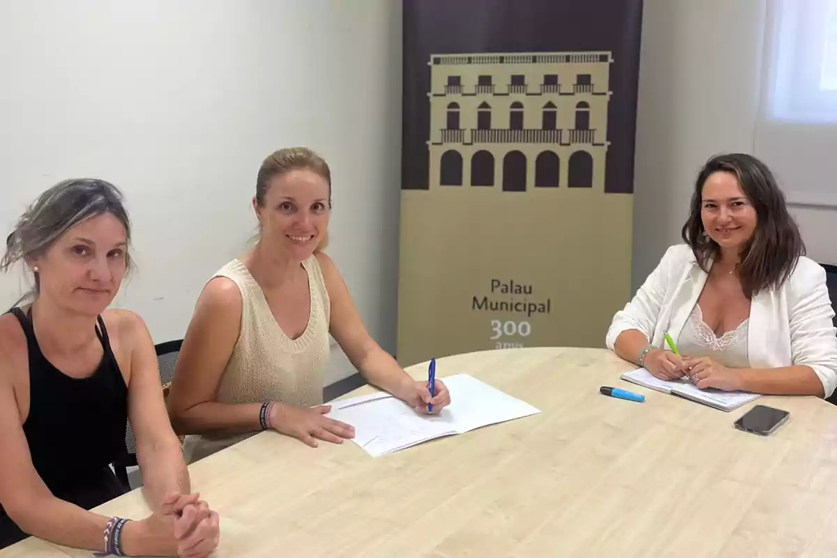 Tres mujeres sentadas alrededor de una mesa de madera en una sala de reuniones, dos de ellas escribiendo en papeles y una pancarta de fondo con el dibujo de un edificio y el texto Palau Municipal 300 anys