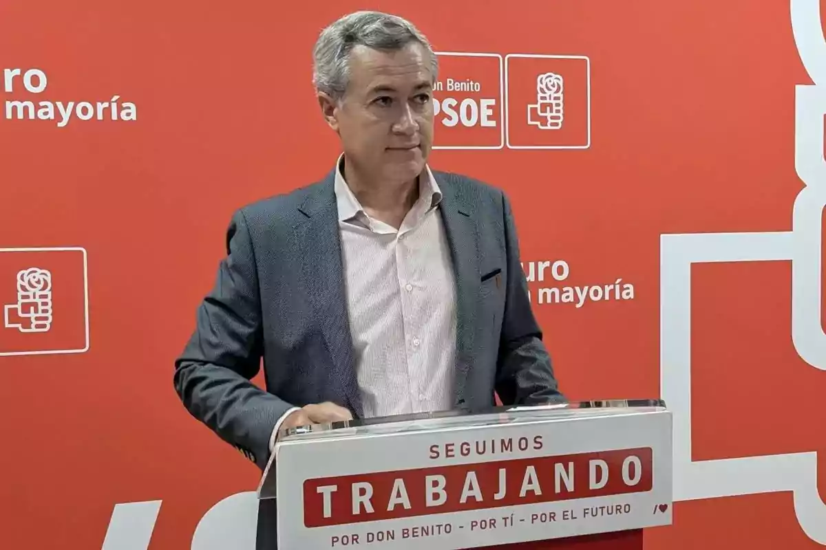 Hombre de cabello canoso con saco gris y camisa clara de pie tras un atril con el texto seguimos trabajando por Don Benito por ti por el futuro sobre un fondo rojo con logotipos y letras del PSOE. Hombre de cabello canoso con saco gris y camisa clara de pie tras un atril con el texto seguimos trabajando por Don Benito por ti por el futuro sobre un fondo rojo con logotipos y letras del PSOE.
