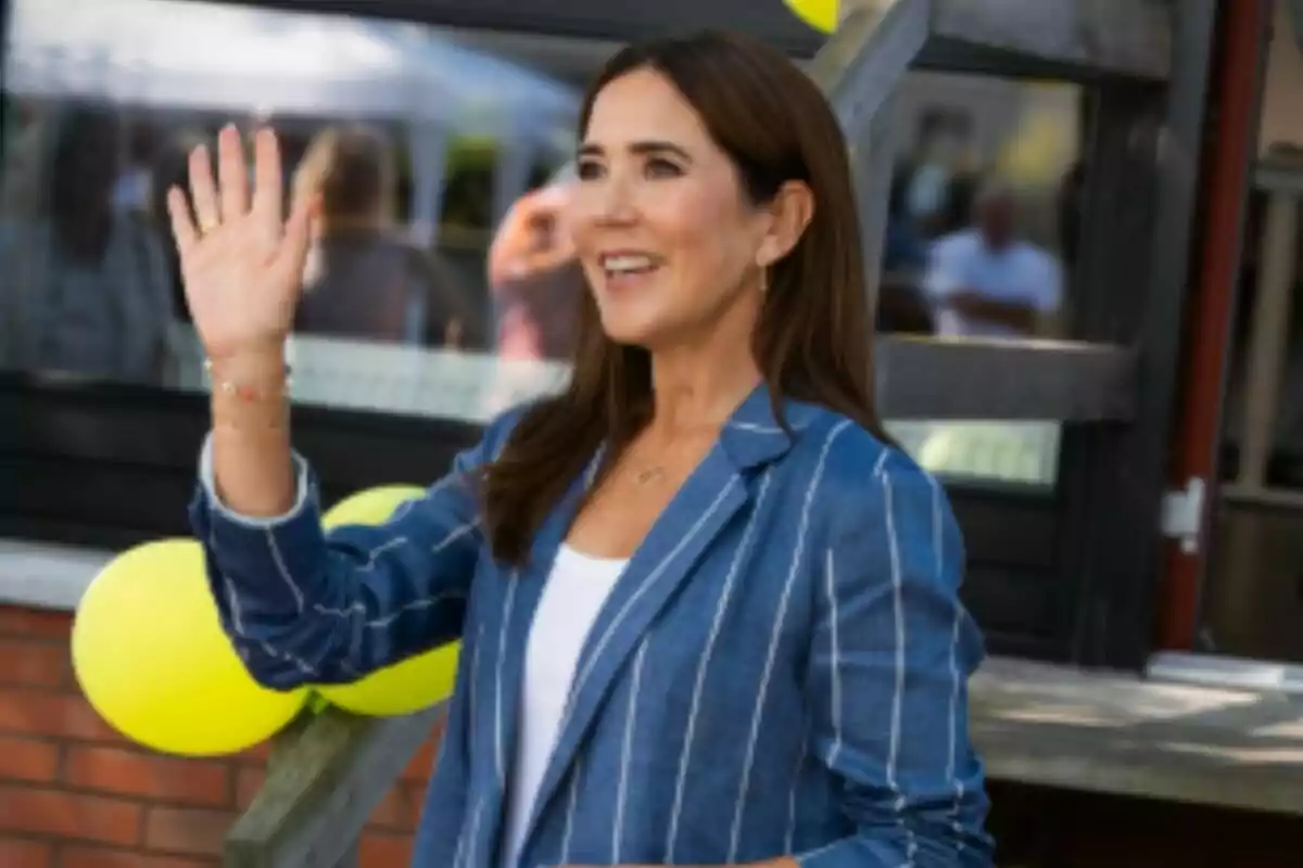 Plano medio de la reina Mary de Dinamarca sonriente con chaqueta azul de rayas saludando con la mano levantada frente a globos amarillos. Plano medio de la reina Mary de Dinamarca sonriente con chaqueta azul de rayas saludando con la mano levantada frente a globos amarillos.