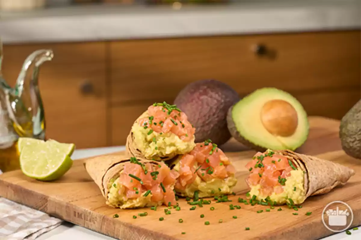 Conos de tortilla rellenos de guacamole y salmón picado decorados con cebollino sobre una tabla de madera con aguacate y lima al fondo Conos de tortilla rellenos de guacamole y salmón picado decorados con cebollino sobre una tabla de madera con aguacate y lima al fondo