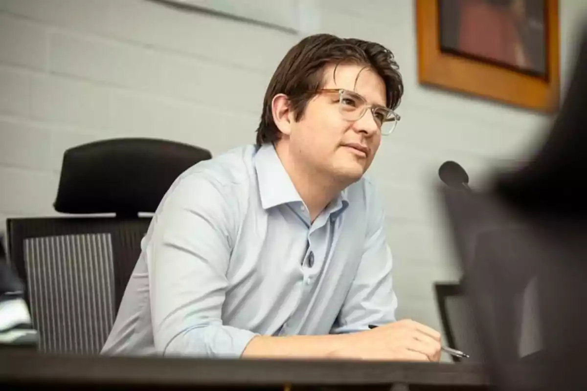Hombre joven con gafas y camisa clara sentado en una silla de oficina mirando hacia el frente con expresión atenta
