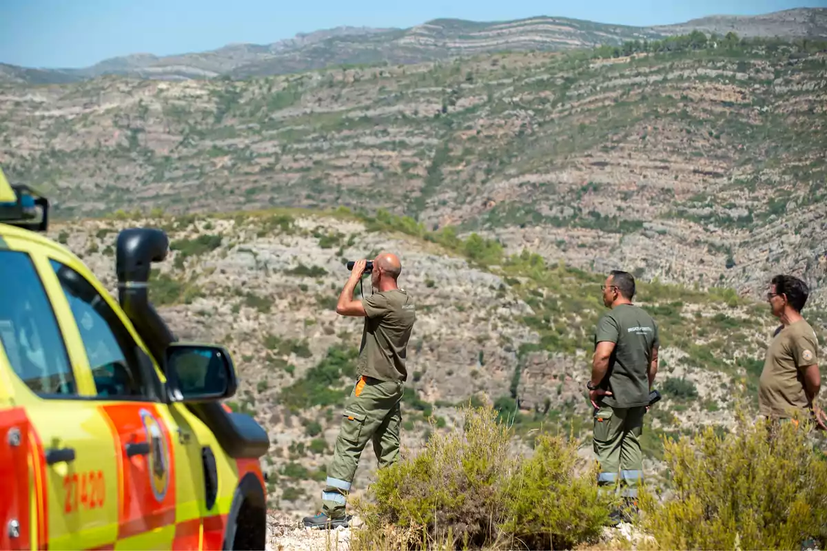 Tres personas con uniforme observan el paisaje montañoso mientras una de ellas usa binoculares junto a un vehículo de emergencia amarillo y rojo
