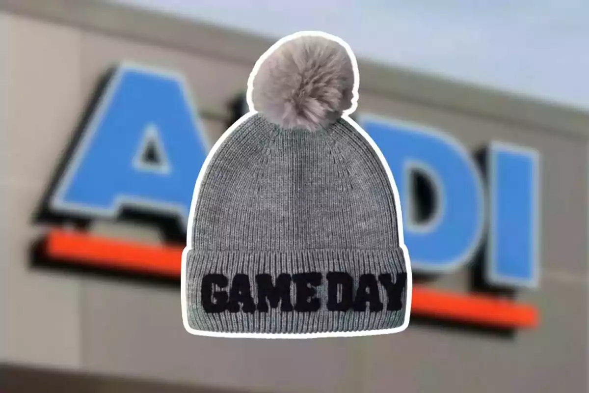 Gorro gris con borla y la frase GAME DAY al frente con el logo de una tienda desenfocado de fondo Gorro gris con borla y la frase GAME DAY al frente con el logo de una tienda desenfocado de fondo
