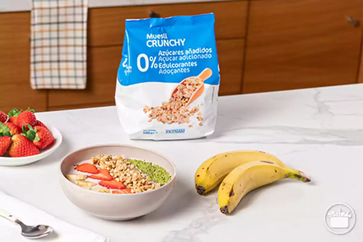 Bol de desayuno con muesli, fresas, plátano y semillas junto a dos plátanos, un plato de fresas y un paquete de muesli crunchy sin azúcares añadidos sobre una encimera blanca Bol de desayuno con muesli, fresas, plátano y semillas junto a dos plátanos, un plato de fresas y un paquete de muesli crunchy sin azúcares añadidos sobre una encimera blanca