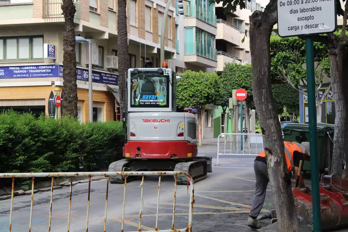 Excavadora Takeuchi trabajando en una calle urbana con un trabajador usando chaleco naranja y señales de tráfico alrededor