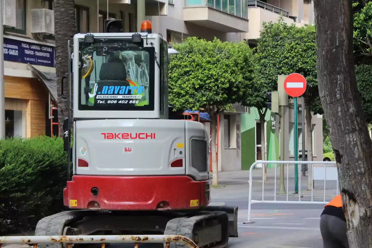 Excavadora Takeuchi estacionada en una calle urbana con árboles y una señal de tráfico de sentido prohibido al fondo Excavadora Takeuchi estacionada en una calle urbana con árboles y una señal de tráfico de sentido prohibido al fondo