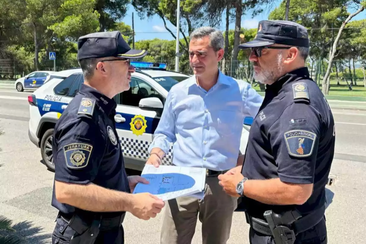 Tres hombres conversan junto a un coche de policía en una calle, dos de ellos llevan uniforme policial y el otro viste camisa clara