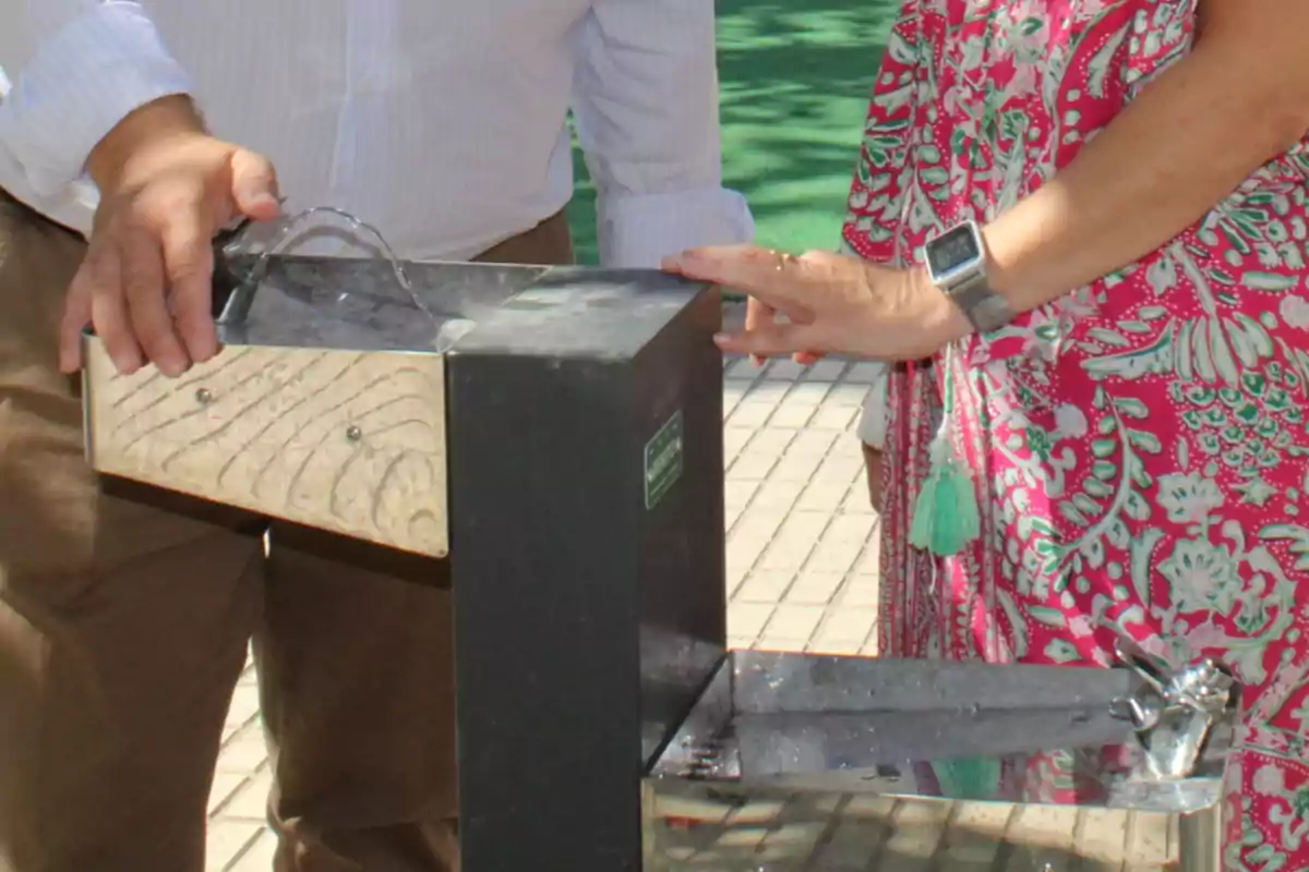 Dos personas usando un bebedero público en un espacio exterior pavimentado Dos personas usando un bebedero público en un espacio exterior pavimentado