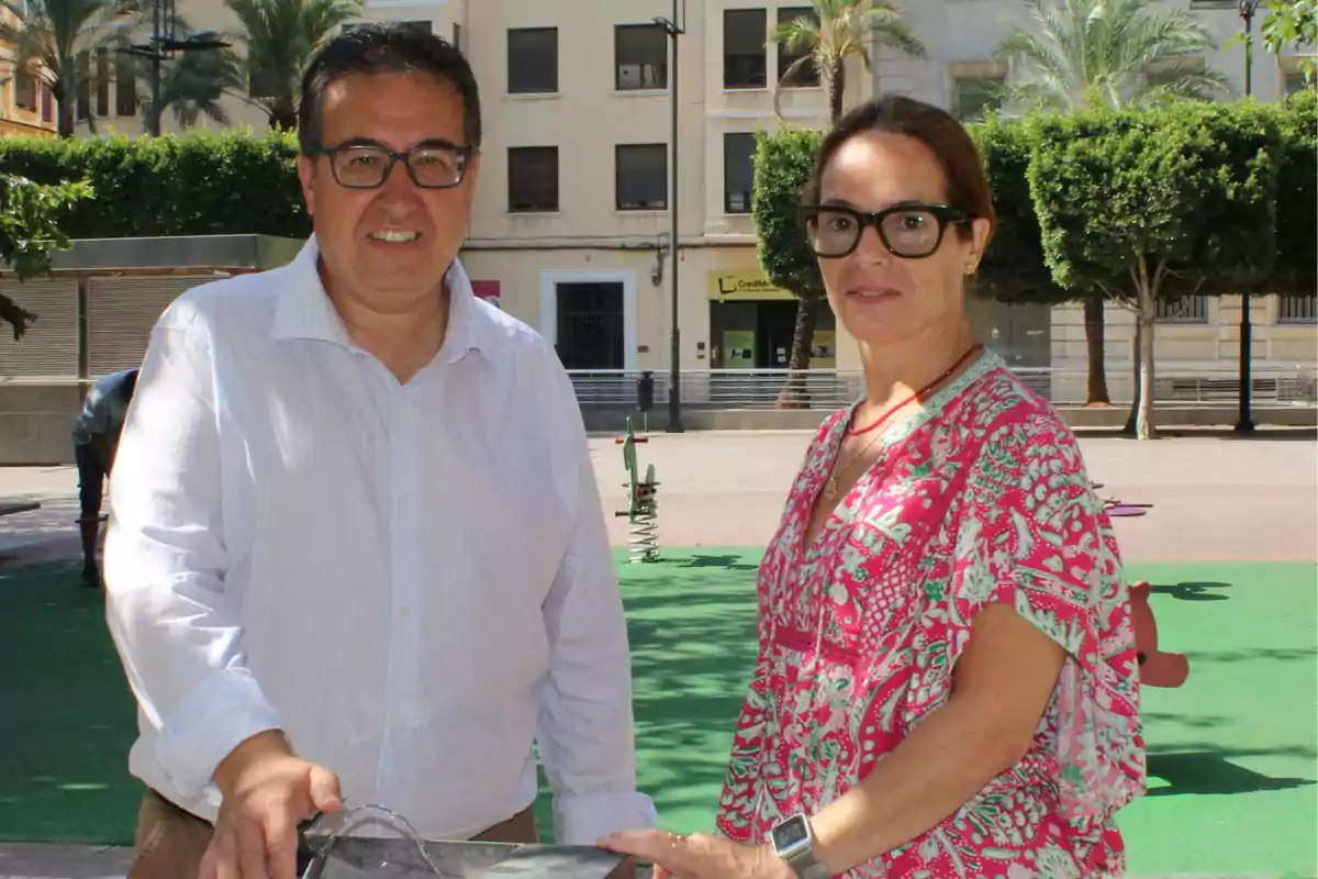 Dos personas con gafas posan sonrientes en un parque urbano con árboles y edificios al fondo Dos personas con gafas posan sonrientes en un parque urbano con árboles y edificios al fondo