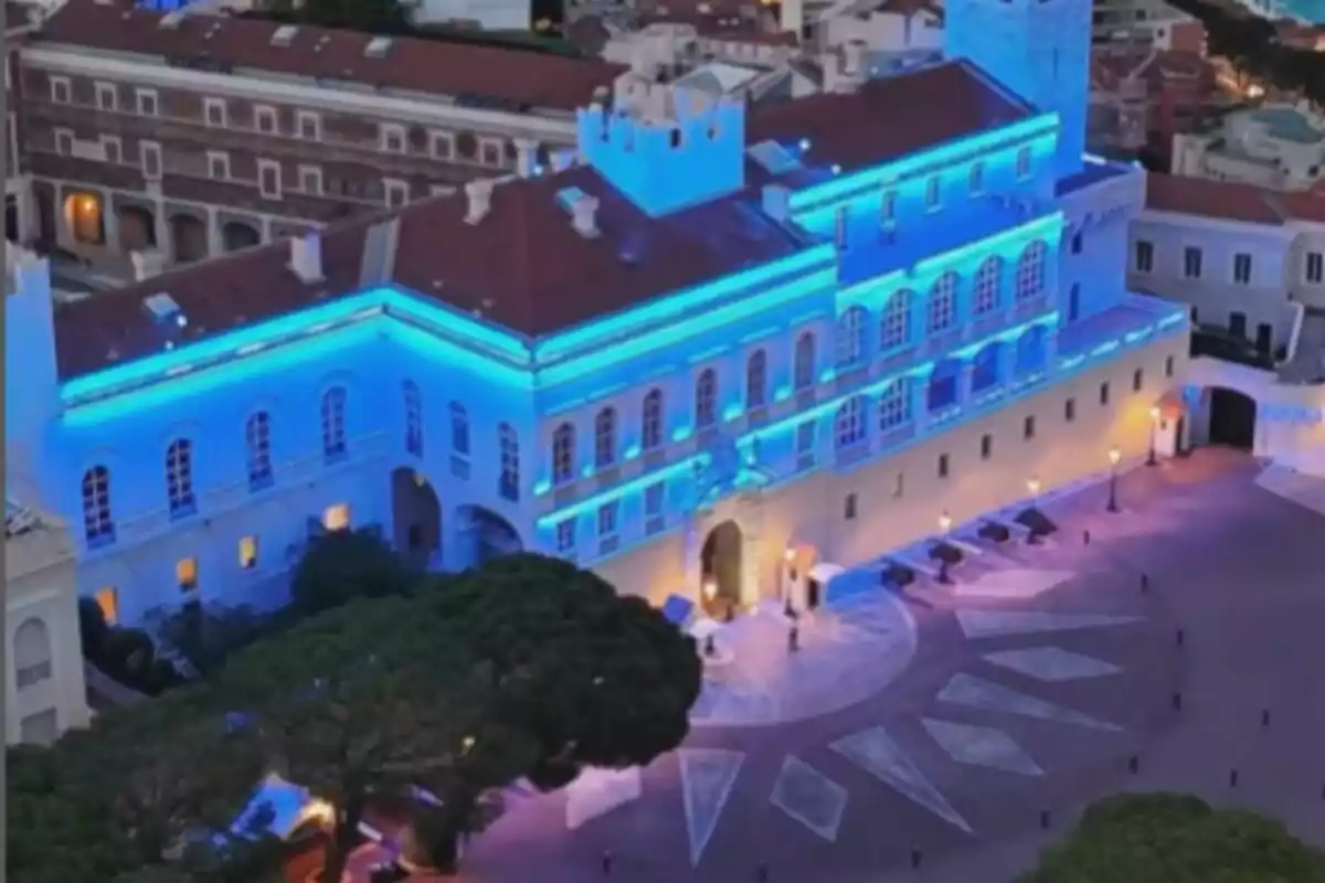 Edificio histórico iluminado con luces azules durante la noche con una plaza amplia al frente y árboles alrededor Edificio histórico iluminado con luces azules durante la noche con una plaza amplia al frente y árboles alrededor
