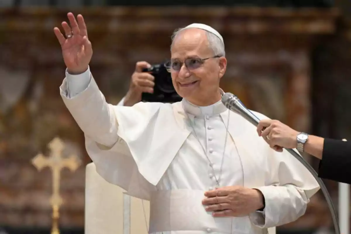 Hombre vestido con vestimenta papal blanca saludando con la mano mientras sonríe y sostiene un micrófono frente a él Hombre vestido con vestimenta papal blanca saludando con la mano mientras sonríe y sostiene un micrófono frente a él