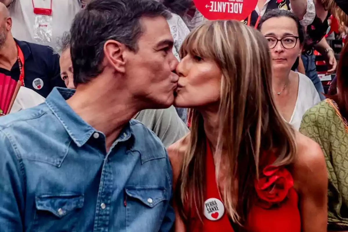 Una pareja se besa en un evento rodeados de personas, ambos llevan ropa casual y hay ambiente festivo. Una pareja se besa en un evento rodeados de personas, ambos llevan ropa casual y hay ambiente festivo.