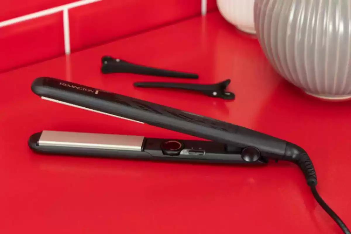 Plancha de cabello negra sobre una superficie roja junto a dos pinzas y un jarrón decorativo Plancha de cabello negra sobre una superficie roja junto a dos pinzas y un jarrón decorativo