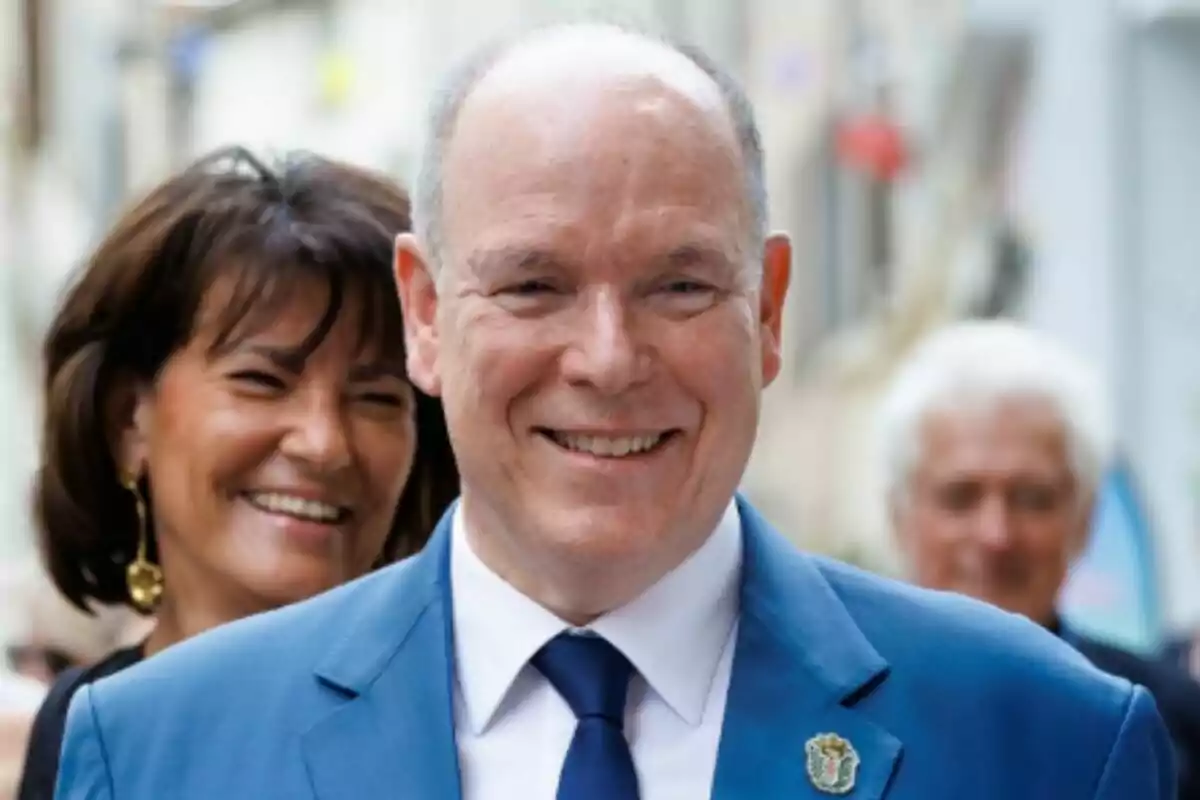 Prince Albert of Monaco dressed in a blue suit smiling while a smiling woman stands behind him in a blurred urban setting. Prince Albert of Monaco dressed in a blue suit smiling while a smiling woman stands behind him in a blurred urban setting.