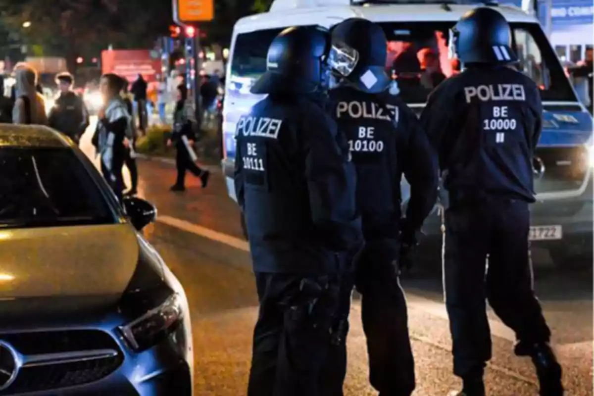 Policías con equipo antidisturbios conversan en una calle por la noche mientras varias personas caminan cerca y hay vehículos estacionados Policías con equipo antidisturbios conversan en una calle por la noche mientras varias personas caminan cerca y hay vehículos estacionados
