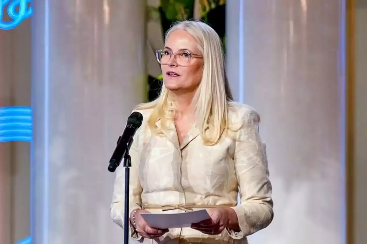 Mette-Marit con gafas hablando frente a un micrófono mientras sostiene unas hojas Mette-Marit con gafas hablando frente a un micrófono mientras sostiene unas hojas