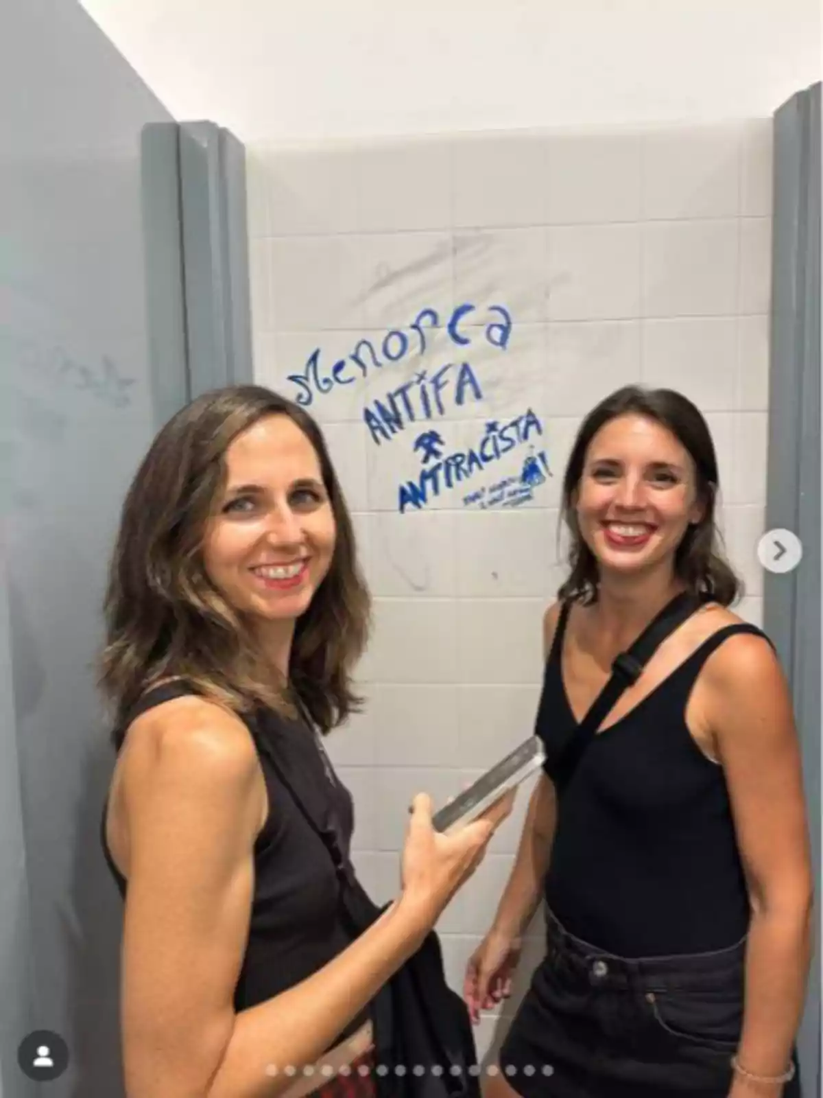 Dos mujeres sonríen en un baño público con paredes blancas, una sostiene un teléfono móvil y detrás de ellas hay un grafiti azul que dice Menorca antifa antirracista. Dos mujeres sonríen en un baño público con paredes blancas, una sostiene un teléfono móvil y detrás de ellas hay un grafiti azul que dice Menorca antifa antirracista.