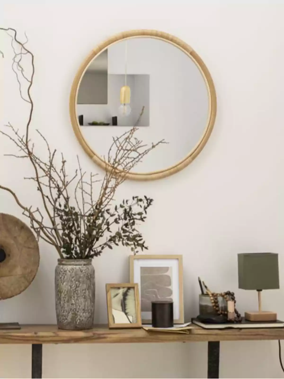 Espejo redondo con marco de madera sobre una mesa de madera decorada con jarrón de ramas secas, cuadros pequeños, una lámpara de mesa verde y varios objetos decorativos Espejo redondo con marco de madera sobre una mesa de madera decorada con jarrón de ramas secas, cuadros pequeños, una lámpara de mesa verde y varios objetos decorativos