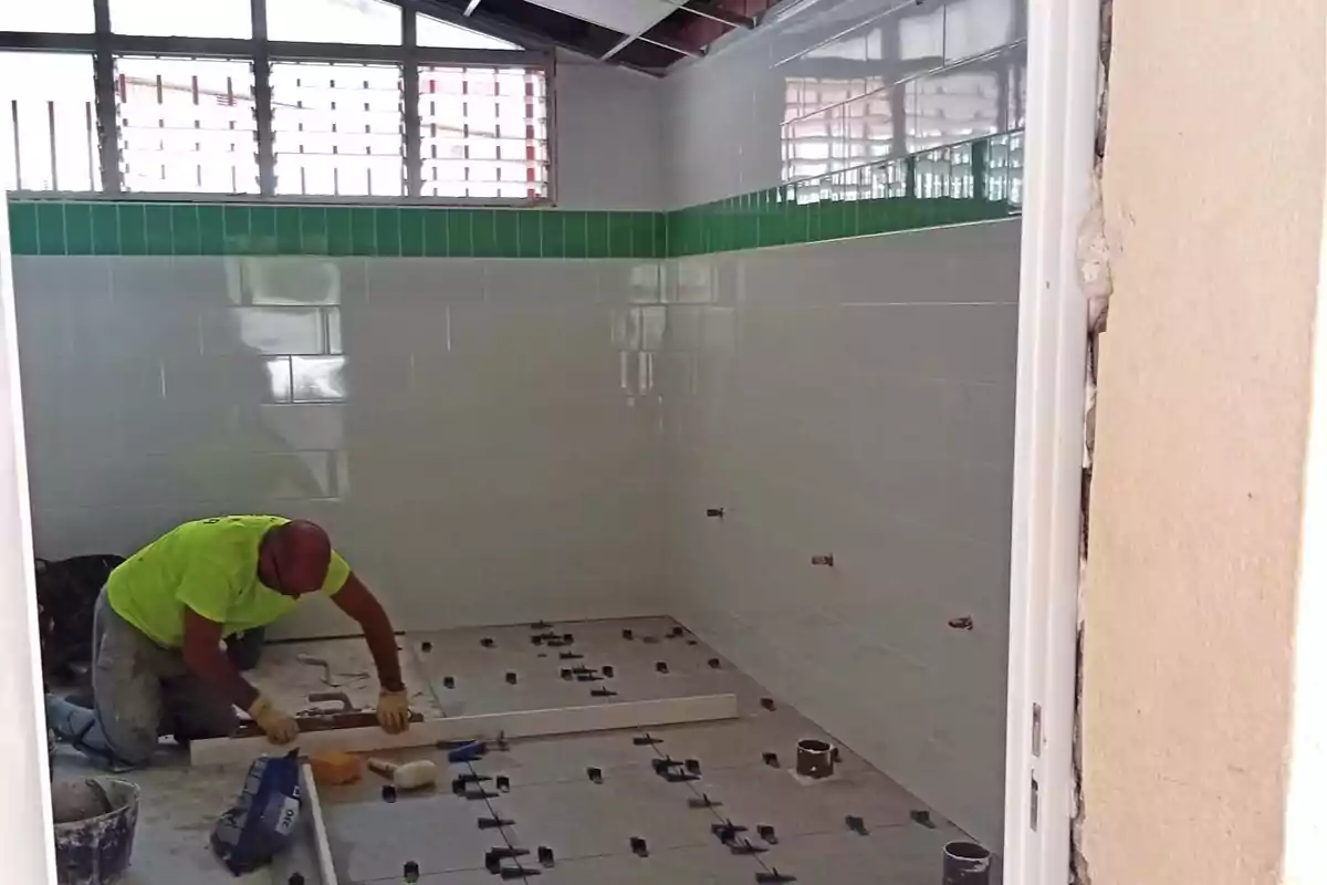 Un hombre instala losas en el piso de una habitación con paredes de azulejo blanco y una franja verde cerca de la ventana. Un hombre instala losas en el piso de una habitación con paredes de azulejo blanco y una franja verde cerca de la ventana.