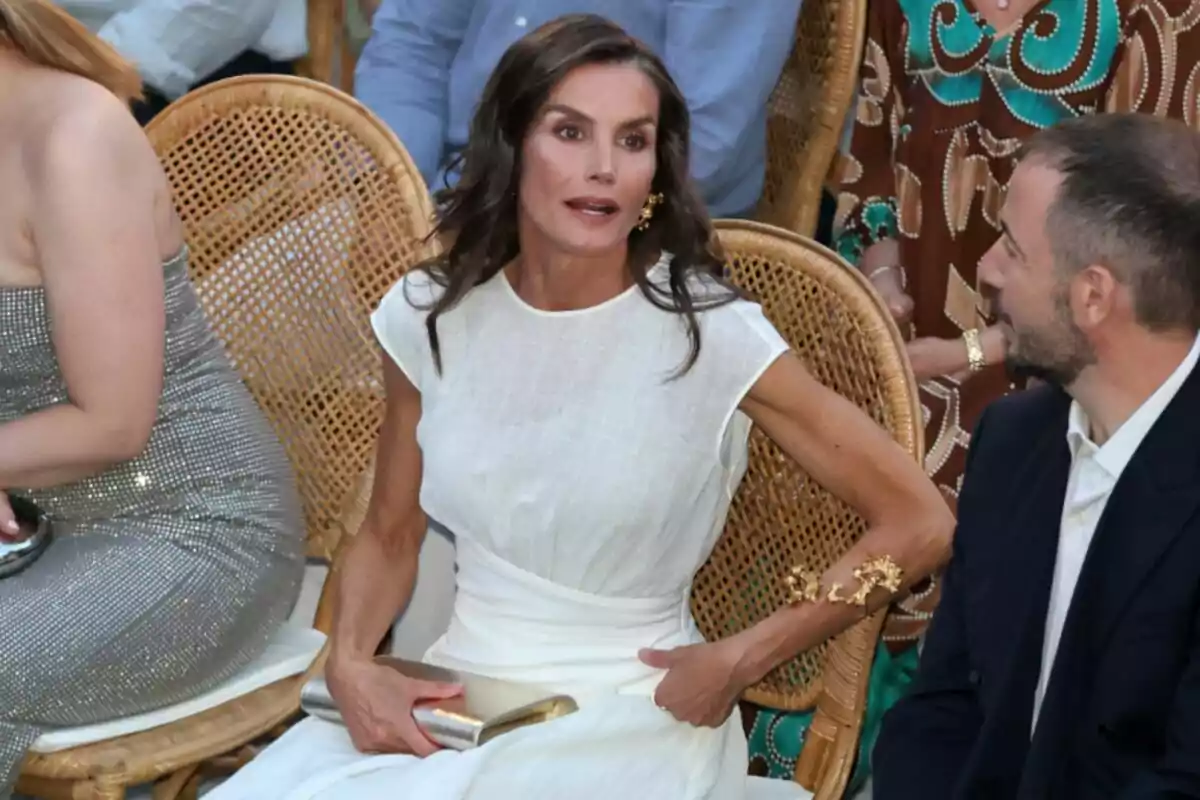 Queen Letizia in a white dress sitting on a wicker chair during a social event, talking with a man