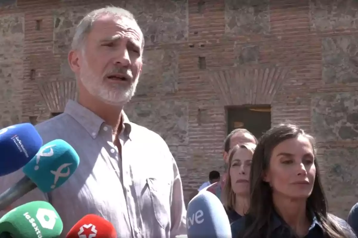 King Felipe, with gray hair and a beard, stands next to Queen Letizia, who has long dark hair, in front of several microphones from different media outlets in an outdoor setting with a brick and stone wall in the background. King Felipe, with gray hair and a beard, stands next to Queen Letizia, who has long dark hair, in front of several microphones from different media outlets in an outdoor setting with a brick and stone wall in the background.