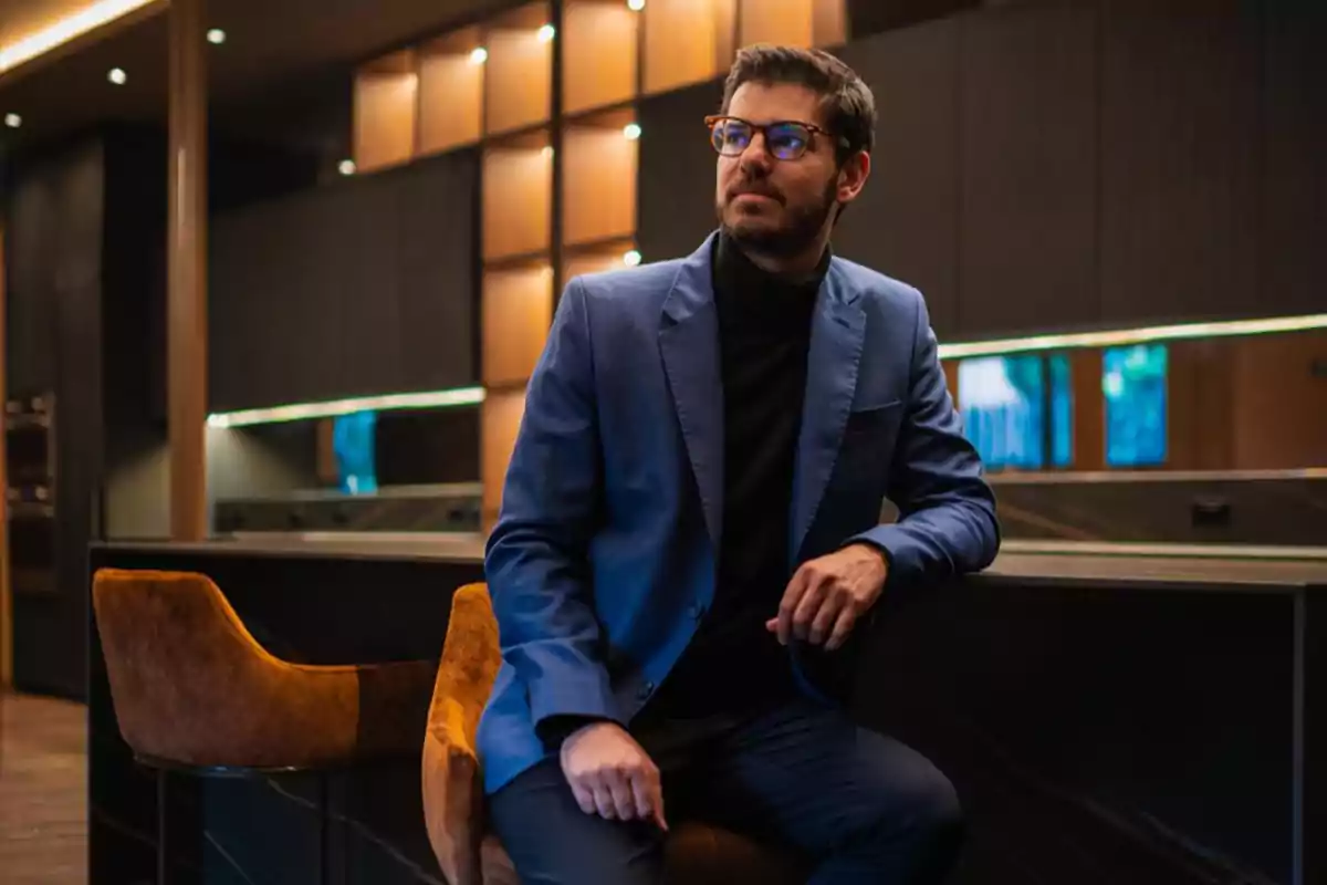 Hombre con gafas y saco azul sentado en una silla en un ambiente elegante y moderno