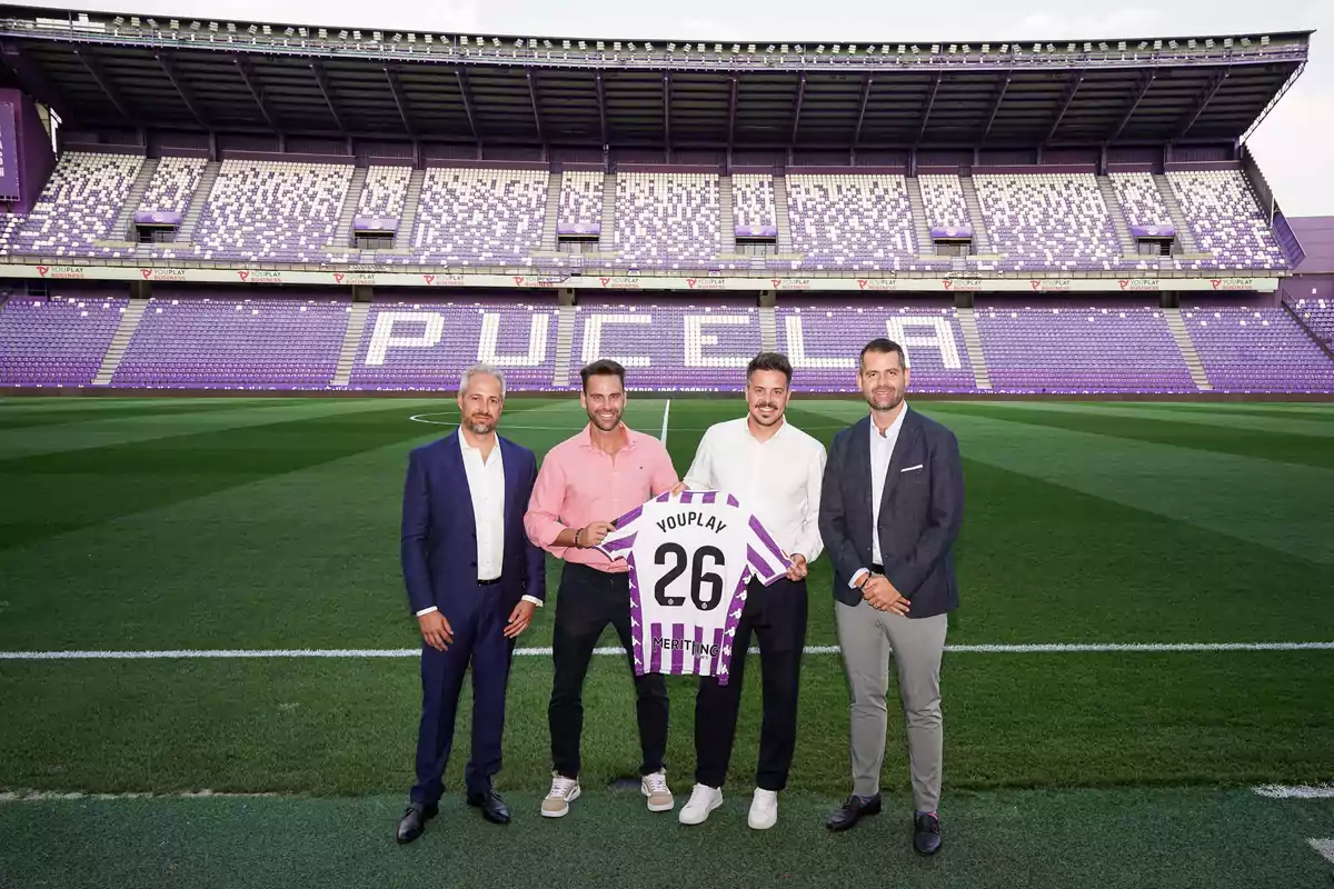 Cuatro hombres posan en el campo de un estadio de fútbol con gradas moradas y blancas que forman la palabra Pucela mientras sostienen una camiseta con el número 26 y el nombre Youplay