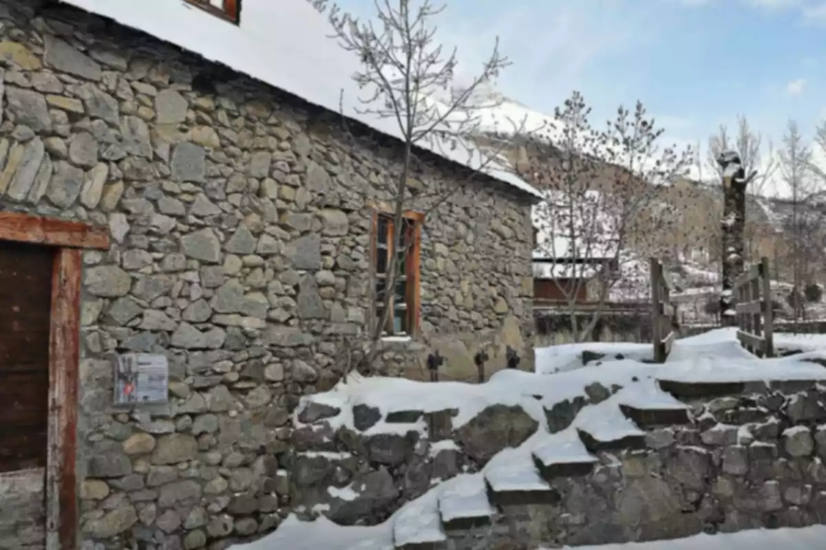 Casa de piedra con techo cubierto de nieve y escaleras exteriores rodeadas de árboles sin hojas en un entorno invernal