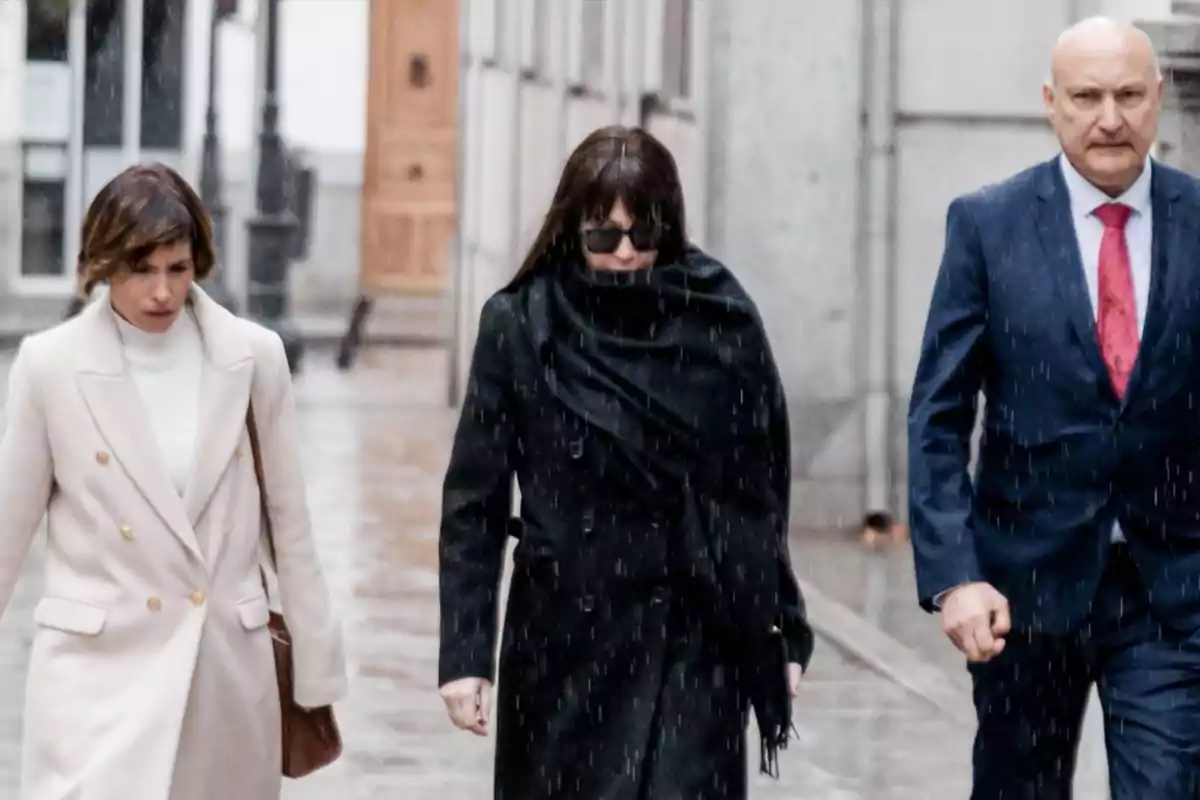Tres personas caminando bajo la lluvia en una calle, una de ellas lleva gafas oscuras y abrigo negro, otra viste un abrigo claro y la tercera lleva traje azul con corbata roja Tres personas caminando bajo la lluvia en una calle, una de ellas lleva gafas oscuras y abrigo negro, otra viste un abrigo claro y la tercera lleva traje azul con corbata roja