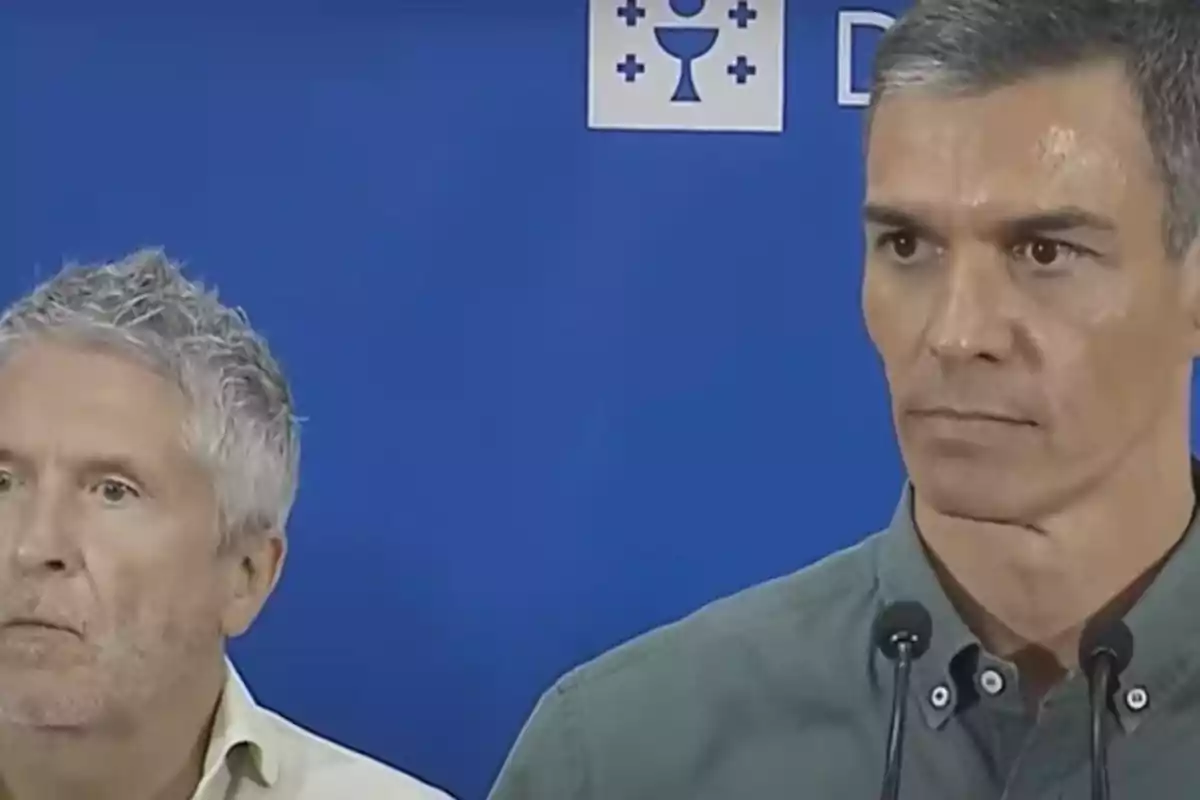 Dos hombres de cabello canoso y expresión seria están frente a un fondo azul con un micrófono y un símbolo institucional Dos hombres de cabello canoso y expresión seria están frente a un fondo azul con un micrófono y un símbolo institucional